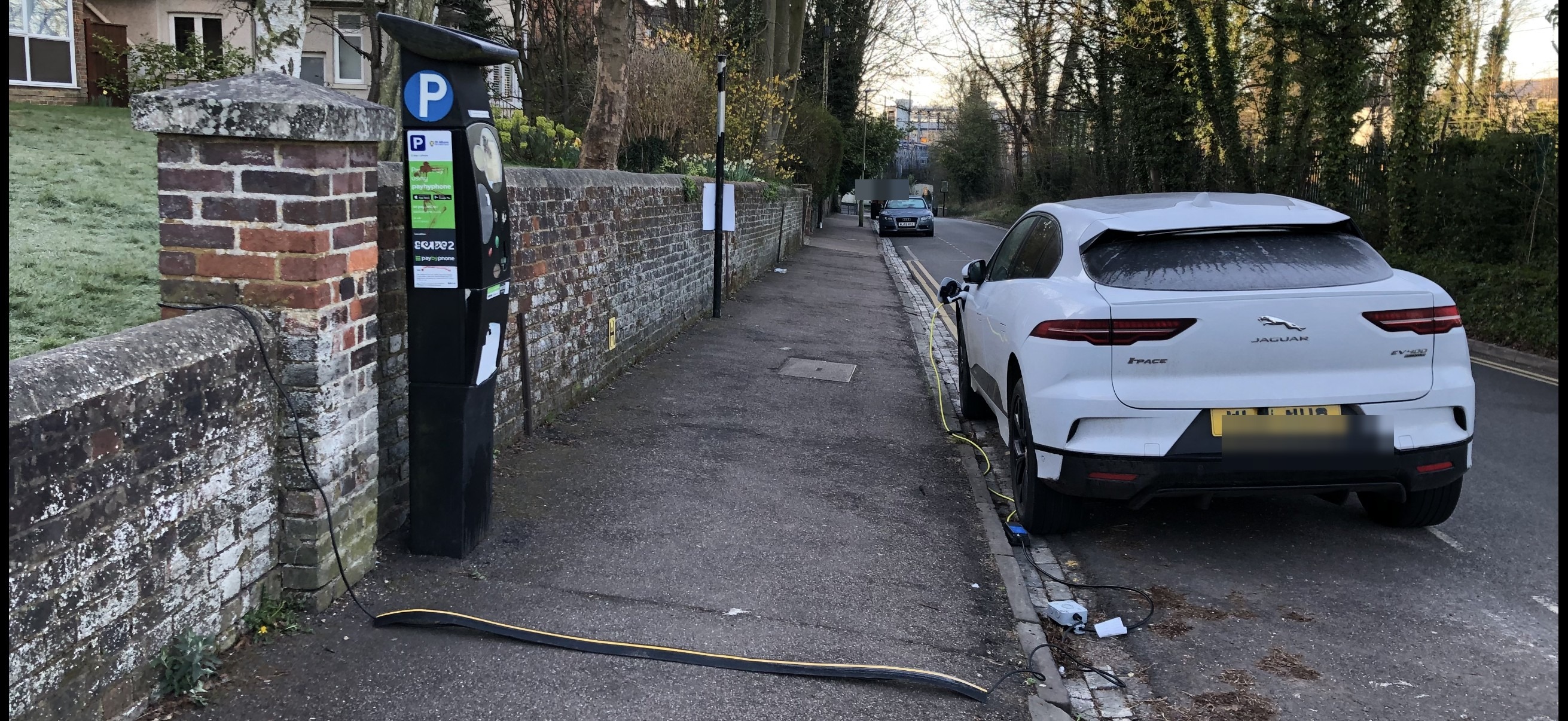 EV charging cable running from an on-street pay-and-display charging point across a UK pavement to a car parked at the kerb, illustrating the lack of off-street parking access