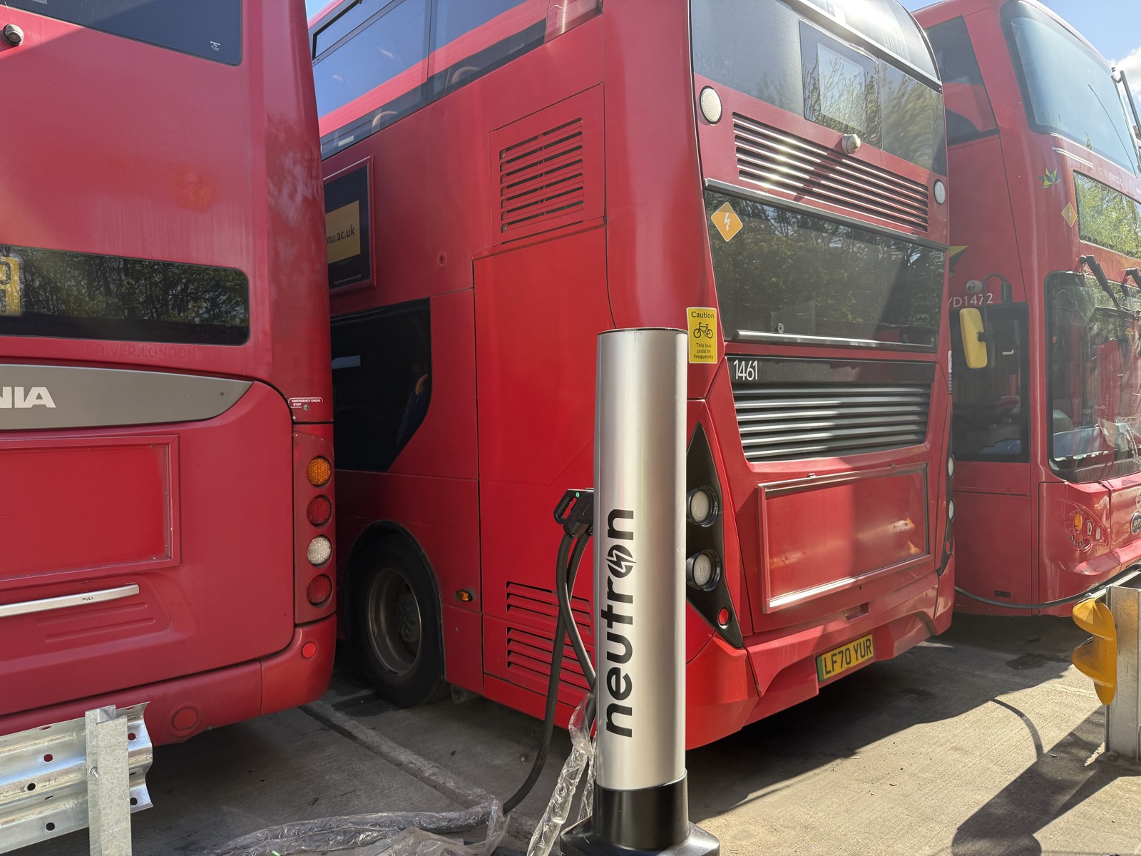 Neutron Systems charging terminals lined up at a UK bus depot alongside a red double-decker bus